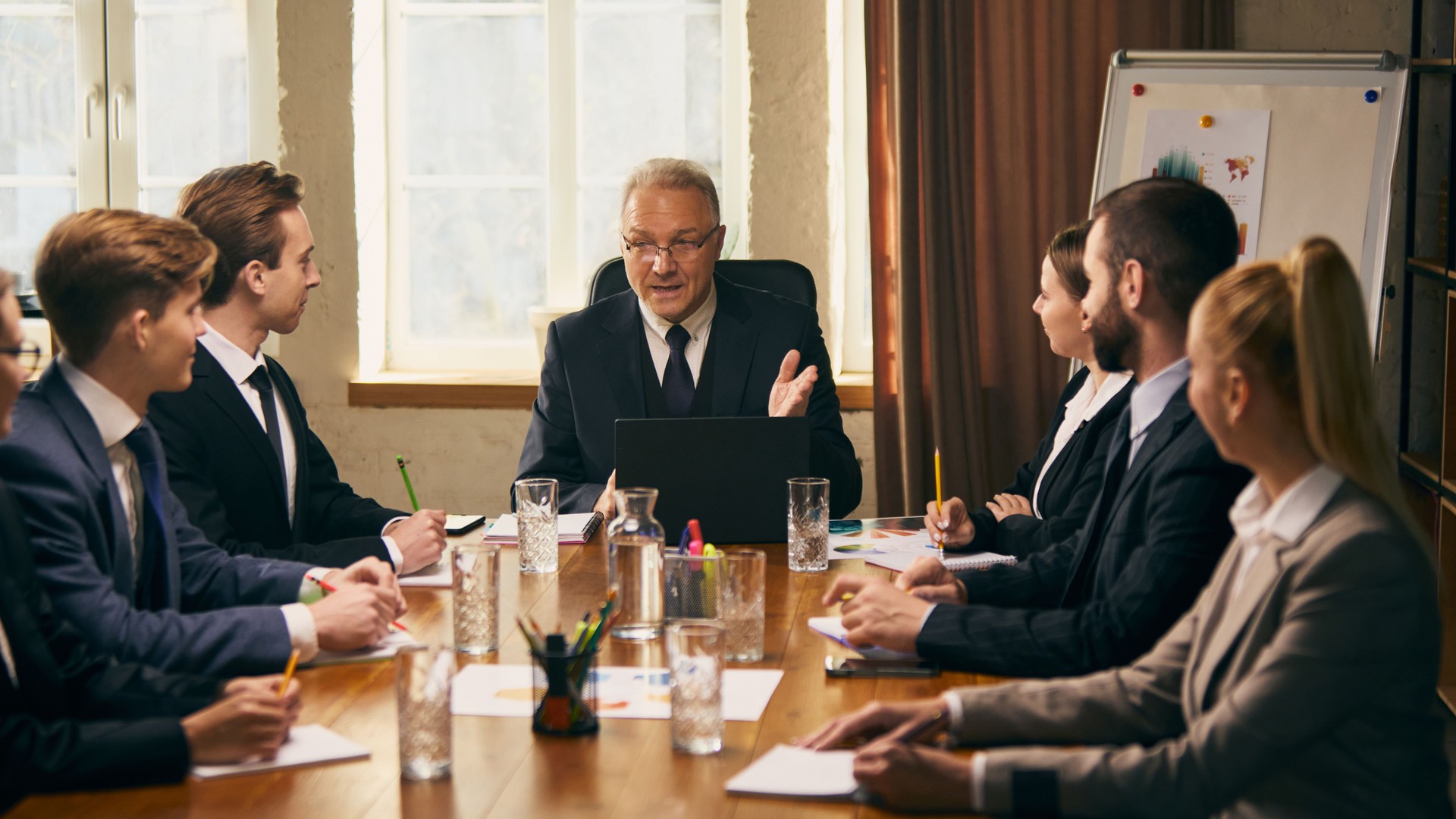 Senior business leader delivers presentation to group of young professionals during team meeting, with graphs and charts visible in background.